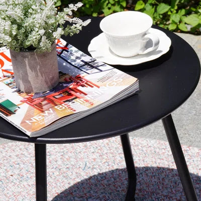 Black round table with a cup, saucer, and magazine on top, outdoors.