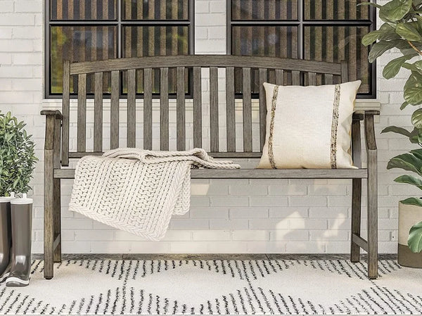 Wooden bench with a blanket and pillow against a white brick wall.